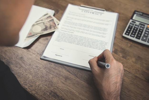 Mano sujetando pluma firmando contrato con dinero, moneda del yen japonés, sobre la mesa