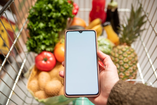 Mano sujetando el teléfono con espacio de copia de pantalla blanca en el carrito de la compra con frutas y verduras alimentos saludables