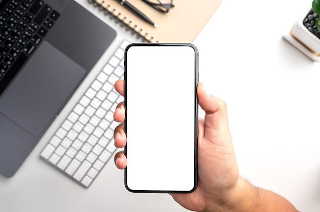 Foto mano sujetando el teléfono inteligente con pantalla en blanco en blanco sobre el escritorio con computadora portátil