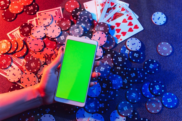 Foto mano sujetando el teléfono con pantalla verde y juego de póquer en la parte posterior de una mesa con luces rojas y azules