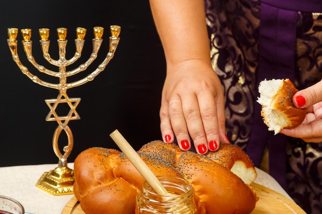Una mano sumerge un trozo de jalá en miel en honor a la celebración de Rosh Hashaná cerca de la mesa miel y jalá y comida tradicional