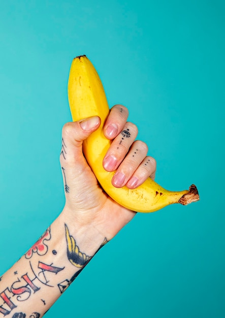 Mano tatuada con un plátano maduro sobre fondo azul.