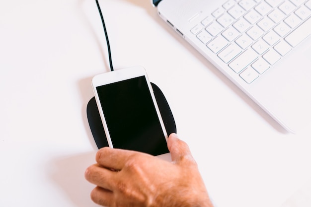 Mano con teléfono inteligente móvil blanco, cargando en una base de cargador inalámbrico junto a una computadora portátil en una mesa de trabajo blanca