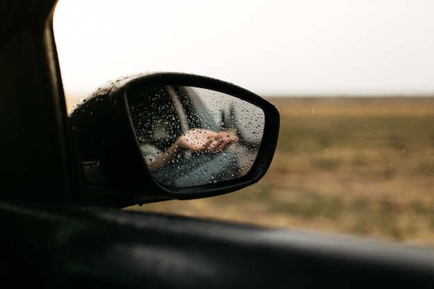 Mano tocando gotas de lluvia. Espejo visto a través del cristal. Ventana de coche mojada. Ciérrese encima de la gota de lluvia. Vista del coche ver el espejo. Día lluvioso.