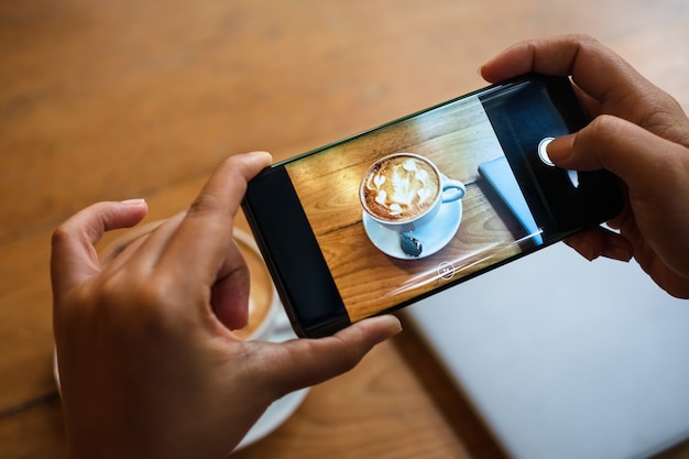 Mano tomando una foto de una taza de café con leche caliente con espuma de arte en la mesa de madera