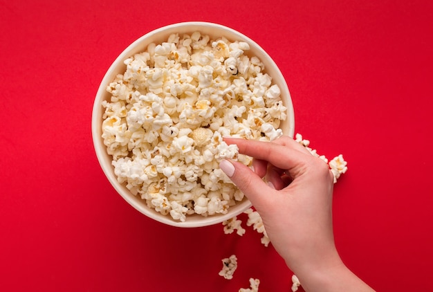 Mano tomando palomitas de maíz del clásico cubo de rayas sobre fondo rojo. Maíz caliente esparcido desde la caja de papel, copie el espacio. Bocadillo de comida rápida y película, vista superior