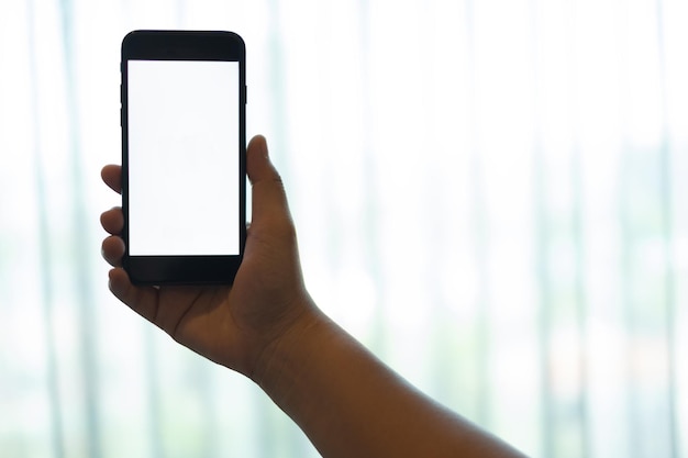 Mano usando un teléfono móvil con pantalla blanca en blanco sobre fondo de cortina blanca