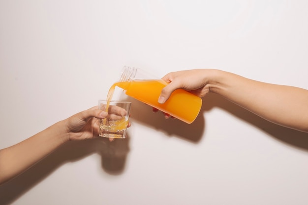 Mano vertiendo jugo de naranja de una jarra con hielo en un vaso aislado sobre fondo blanco.