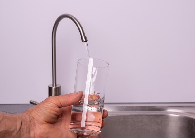 Foto mano vertiendo un vaso de agua del grifo del filtro, pared blanca, espacio de copia