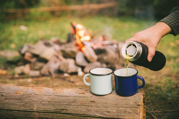 Mano vierta té en tazas de metal fuego de campamento en el fondo