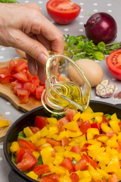 Foto mano vierte aceite. tomates en rodajas y verduras en una sartén. huevos, aceitunas, ajo, pimiento amarillo en la mesa.