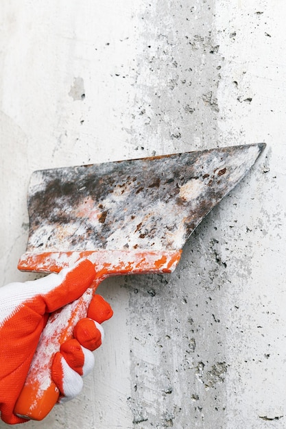 La mano del yesero sostiene un viejo cuchillo de masilla de hoja flexible y raspa la pared de concreto quita el yeso ...