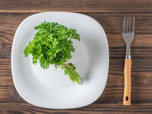 Un manojo de perejil fresco en un plato blanco y un tenedor sobre una mesa de madera