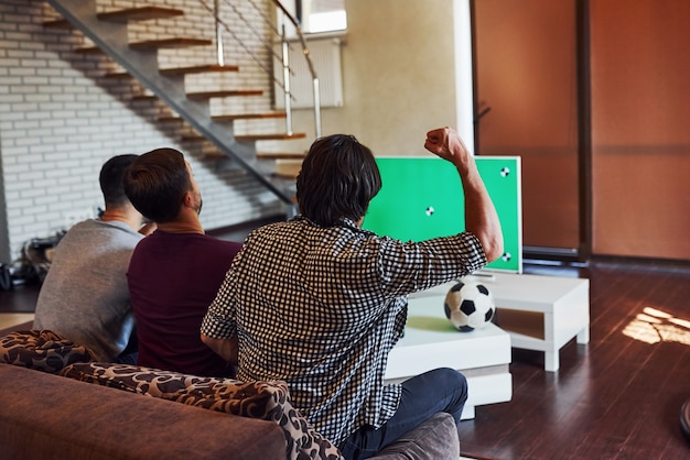 Con las manos en alto. Emocionados tres amigos viendo fútbol en la televisión en casa juntos.