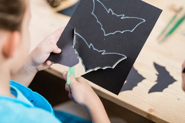 Manos de alumno elementrary cortando murciélago de halloween de papel negro mientras está sentado junto al escritorio de madera en la lección
