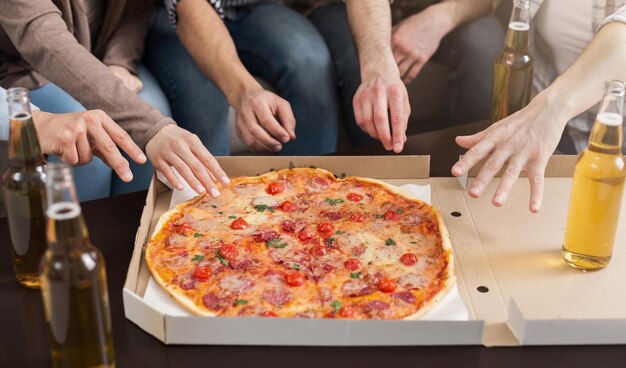 Manos de amigos tomando porciones de pizza sabrosa y bebiendo de botellas en casa