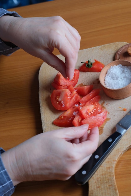 Foto manos de una anciana cortando un tomate rojo maduro en una tabla de cortar para tomar un refrigerio en la cocina en una mesa marrón sin rostro cerrado