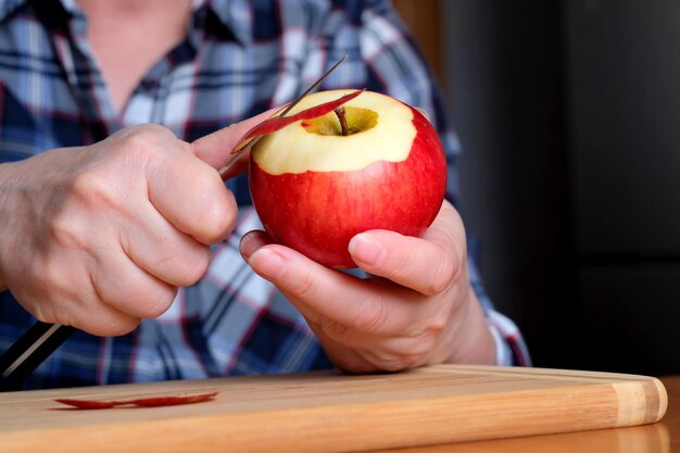 Las manos de una anciana pelan una manzana jugosa, madura y roja con un cuchillo en la cocina en una mesa marrón sin cara cerrada