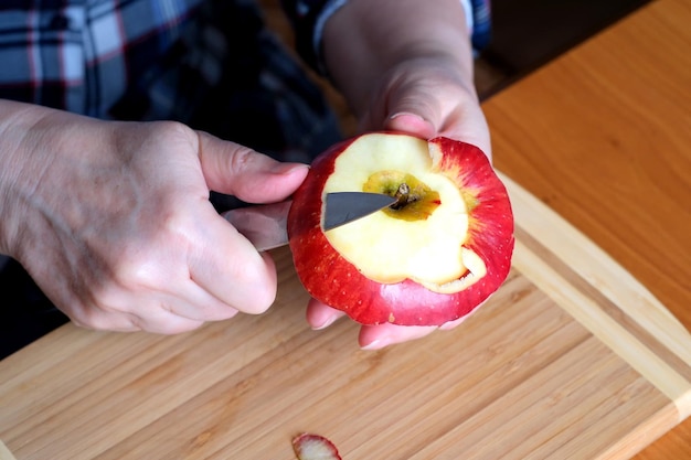 Las manos de una anciana pelan una manzana jugosa, madura y roja con un cuchillo en la cocina en una mesa marrón sin cara cerrada