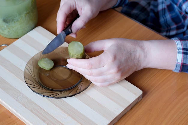 Las manos de una anciana sacan deliciosos pepinos encurtidos caseros de un frasco para cortarlos como refrigerio en la cocina en una mesa marrón sin rostro cerrado