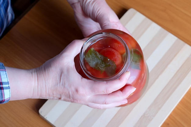 Las manos de una anciana sacan deliciosos tomates encurtidos caseros de un frasco en la cocina en una mesa marrón sin rostro cerrado