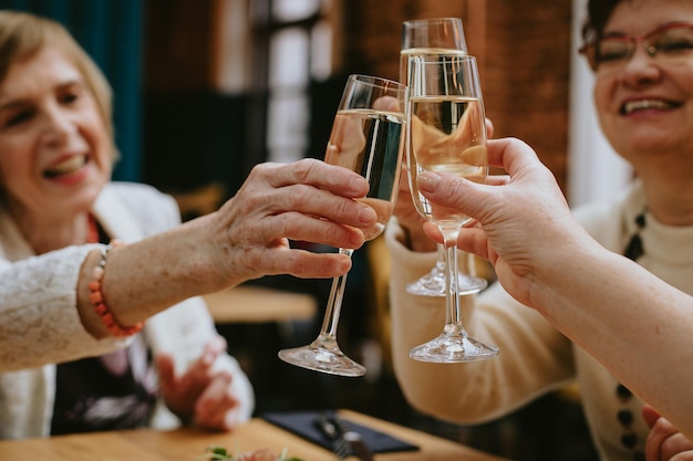 Manos de ancianas sonrientes que tocan copas de vino o champán mientras celebran su reunión o alguna festividad en un acogedor café o restaurante