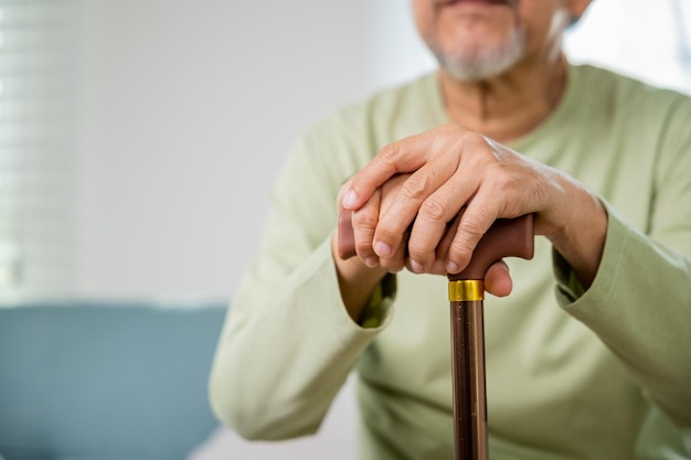 Manos de un anciano asiático sentado descansando en casa sostienen un bastón de madera