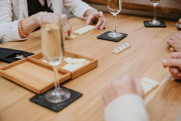 Manos de ancianos irreconocibles posiblemente mujeres jugando dominó en un acogedor café al aire libre acostados en la mesa con copas llenas de champán o vino celebrando su reunión