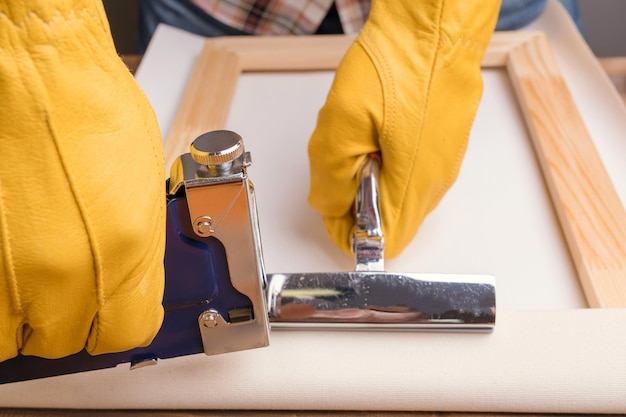 Las manos del artesano con guantes protectores amarillos colocan el lienzo en una camilla de madera con una pistola grapadora y unas pinzas.