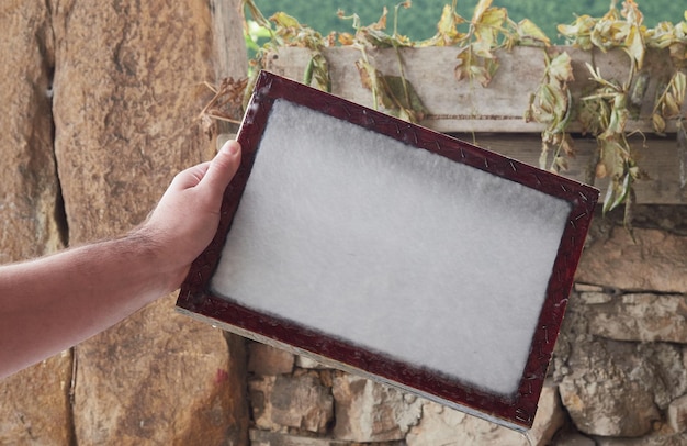 Manos de artesano sosteniendo un marco de madera con fibras vegetales para hacer hojas de papel. Antiguo proceso tradicional de producción de papel.