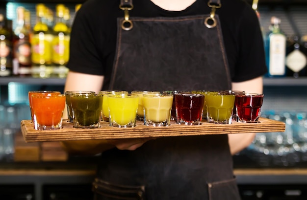 En las manos del barman hay un colorido juego de cócteles alcohólicos en vasos de chupito para una fiesta alcohólica