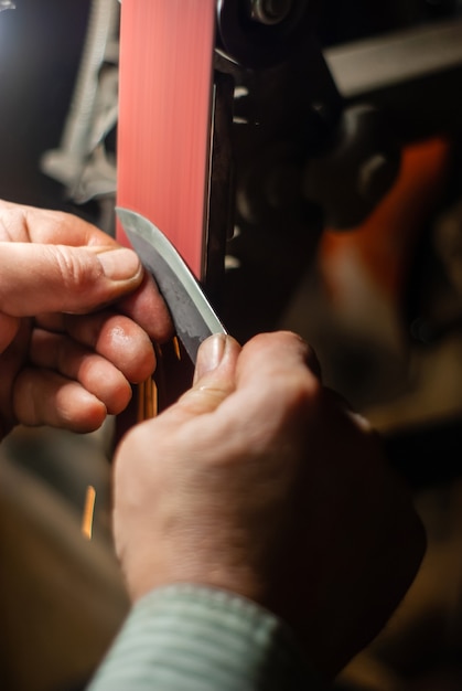 Las manos de un cerrajero afilando la pieza de trabajo para un cuchillo en una cinta abrasiva