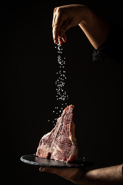 Manos del chef cocinar filete de ternera wagyu porterhouse envejecido en seco con un trozo de filete grande agregando sal y pimienta en un movimiento de congelación sobre una superficie negra