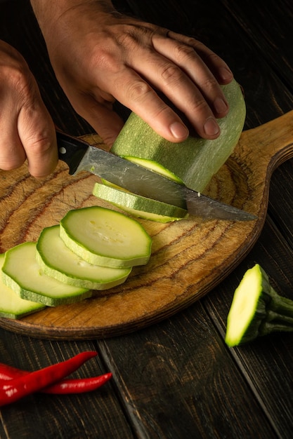 Manos de chef con cuchillo cortando calabacín en tabla de cortar de madera antes de preparar una comida deliciosa