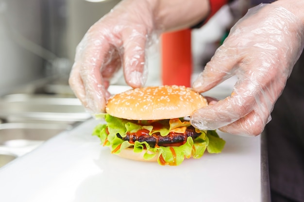 Las manos del chef demuestran la hamburguesa terminada. Preparando hamburguesas en restaurante. Manos del cocinero en guantes desechables según los requisitos de higiene.