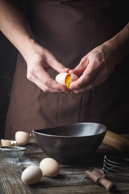 Manos de chef profesional están rompiendo un huevo en un tazón para hacer masa en la mesa de madera, sobre la oscuridad