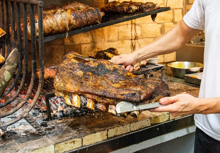 Foto las manos del chef sosteniendo una gran pieza de carne frente a una típica parrilla uruguaya llamada parrilla