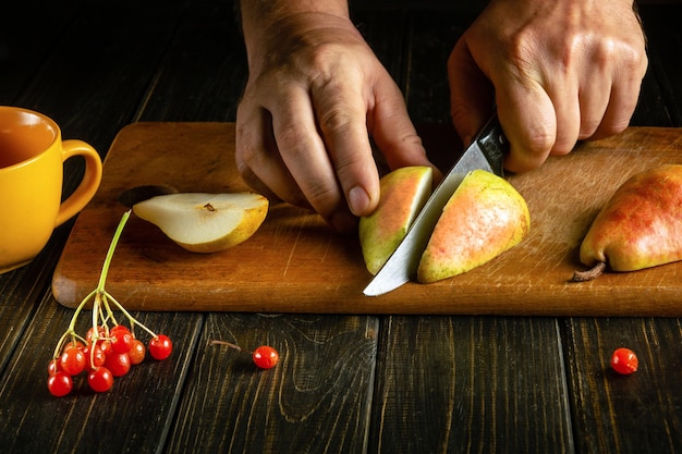 Las manos del chef usan un cuchillo para cortar una pera amarilla en una tabla de cortar para preparar compota o jugo de fruta