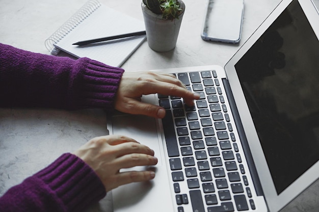 Manos de chicas jóvenes escribiendo en la computadora portátil en el escritorio blanco con planta y cuaderno