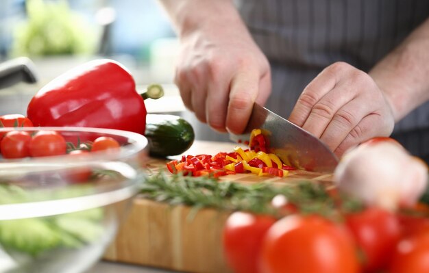 Manos de cocinero cortan pimientos amarillos y rojos en la tabla de cortar con otras verduras en la mesa de la cocina