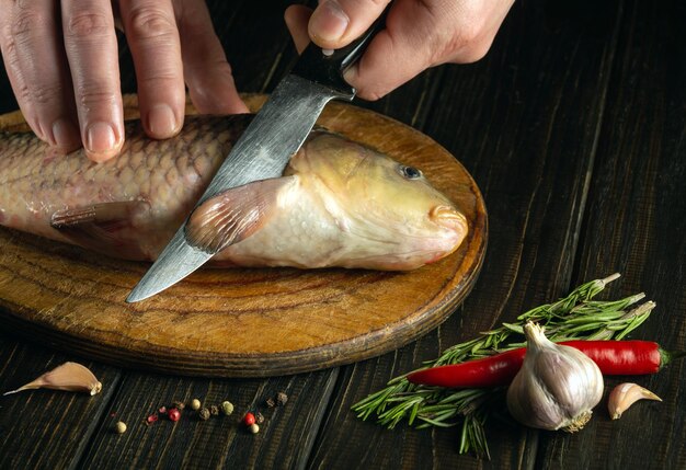 Las manos del cocinero con un cuchillo en la mesa de la cocina antes de limpiar la carpa de pescado cocinando un plato de pescado en la mesa De la cocina con especias y pimienta