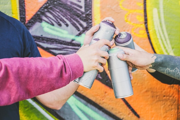 Foto manos cortadas de amigos sosteniendo botellas de pintura en aerosol contra la pared de graffiti