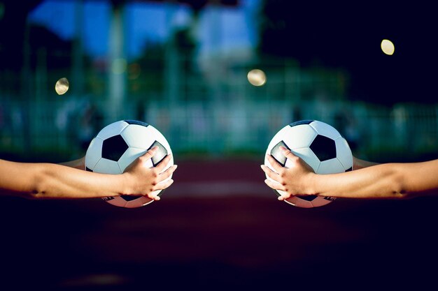 Manos cortadas de personas sosteniendo pelotas de fútbol en la cancha deportiva durante el anochecer