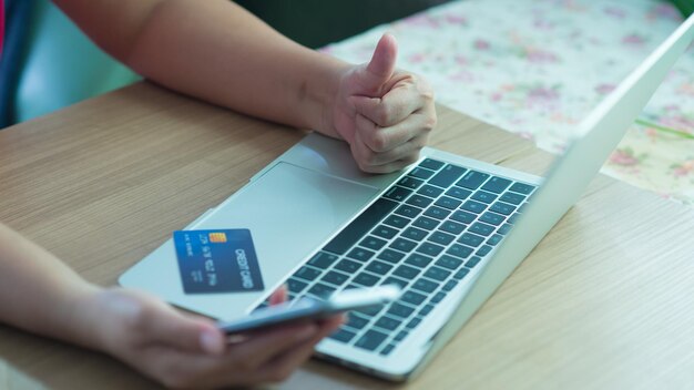 Manos cortadas con tarjeta de crédito usando teléfono y portátil en la mesa