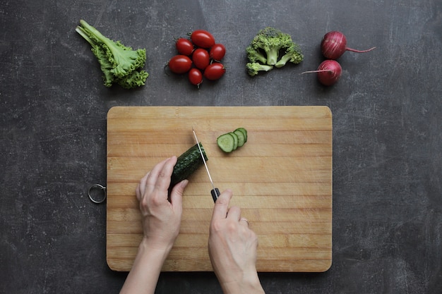 Las manos cortan un pepino en una tabla de cortar sobre una mesa gris.