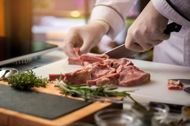 Las manos con cuchillo cortan la carne. Hierbas cerca de la placa de cocción. Chef experto prepara costillas de ternera. Descubra la mejor receta.