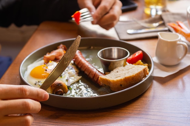 Manos con cuchillo y tenedor y desayuno continental Hombre comiendo huevos fritos con salchichas tomate