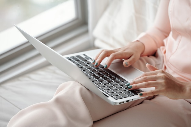 Manos cuidadas de una mujer joven en pijama trabajando en una computadora portátil en casa debido al bloqueo