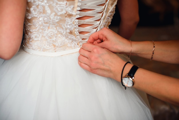 Las manos de la dama de honor abrochan los botones en la parte posterior de la novia en el vestido de novia de seda blanca