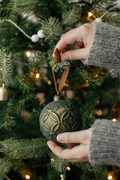 Manos decorando el árbol de Navidad con adornos elegantes en la sala festiva atmosférica Feliz Navidad Preparación para las vacaciones de invierno Mujer en suéter acogedor poniendo juguete vintage en el árbol